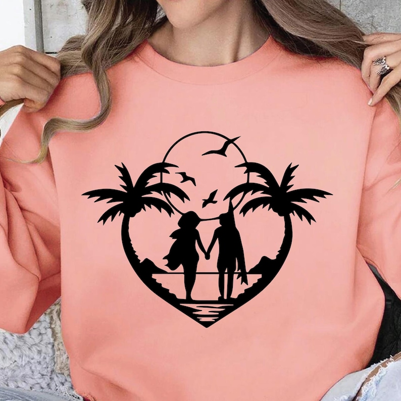 May include: A coral-colored sweatshirt featuring a black silhouette design. The design depicts a couple holding hands on a beach, framed by palm trees and a heart shape. Birds fly in the sky above the couple, with a sun in the background.