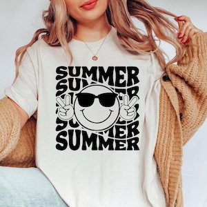 May include: Cream-colored t-shirt with a black graphic design. The design features the word "SUMMER" repeated around a smiling face wearing sunglasses and making a peace sign with both hands. A casual summer tee.