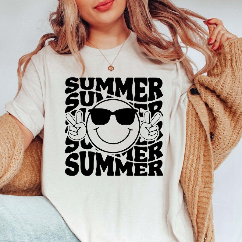 May include: Cream-colored t-shirt with a black graphic design. The design features the word "SUMMER" repeated around a smiling face wearing sunglasses and making a peace sign with both hands. A casual summer tee.