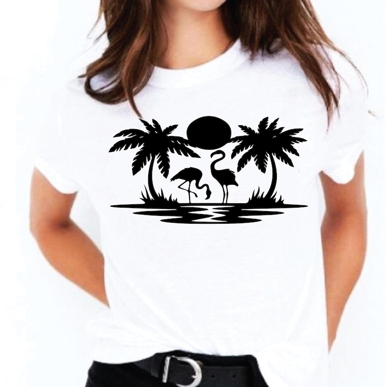 May include: White t-shirt featuring a black graphic of two flamingos, palm trees, and a sun over water. The design is centered on the shirt. The shirt has short sleeves and a crew neck.