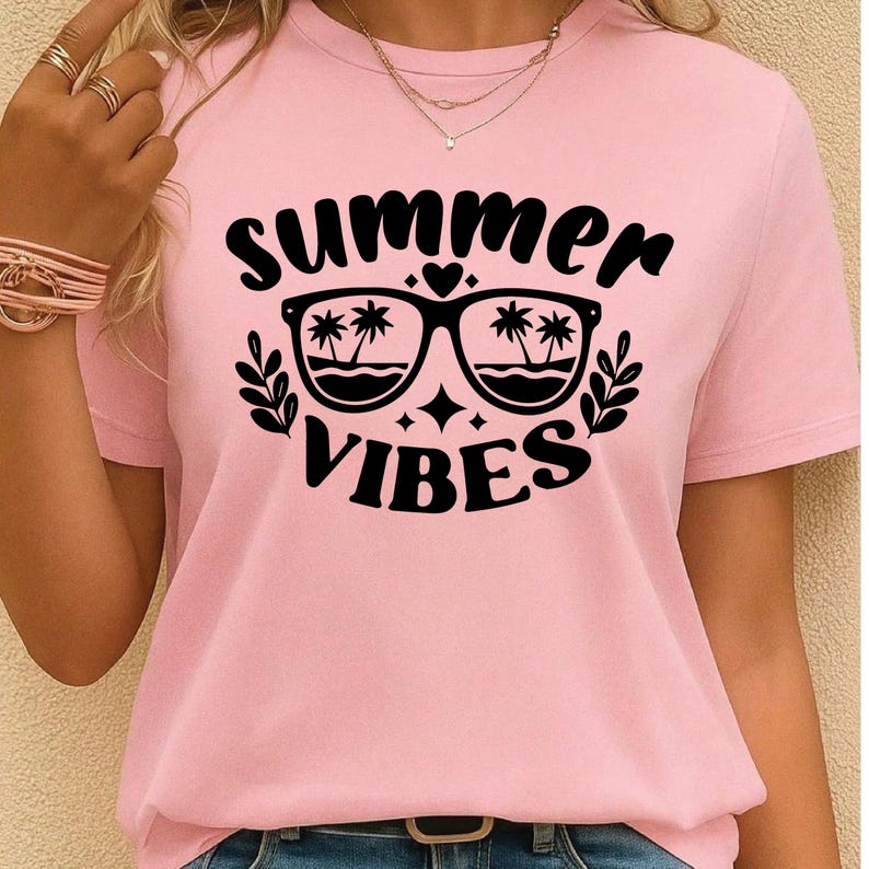 May include: A light pink t-shirt with the words "summer vibes" in black script. The design includes sunglasses with palm trees reflected in the lenses, a heart, and starbursts. The shirt is short-sleeved.