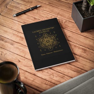 May include: A black journal with gold lettering and geometric designs. The cover reads "SACRED GEOMETRY JOURNAL" with the words "Notes • Patterns • Meditations" below. A black pen and a cup of coffee are also visible.