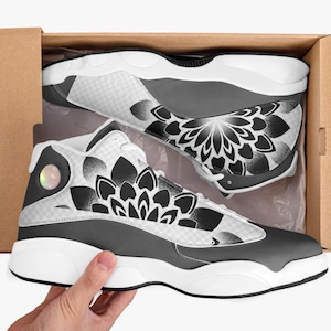 May include: High-top sneakers in a cardboard box. The shoes feature a black, white, and gray color scheme with a floral design on the sides. The soles are white, and the uppers are a mix of gray and white checkered patterns.