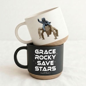 May include: Two ceramic mugs stacked on top of each other. The top mug is white with a brown base and a figurine of a person riding a creature. The bottom mug is black with the words "GRACE ROCKY SAVE STARS" in white.