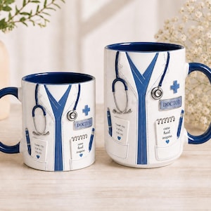 May include: Two white ceramic mugs with blue interiors and handles, designed to resemble a doctor's white coat. Each mug features a stethoscope, a blue cross, a name tag reading DOCTOR, and pockets with notepads printed with text and blue hearts.