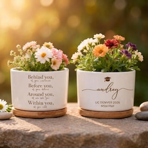 Può includere: Due vasi di fiori in ceramica bianca con sottovasi in legno, ciascuno pieno di fiori colorati. Un vaso ha il testo "Behind you, Before you, Around you, Within you, all you need." L'altro vaso è personalizzato con il nome "audrey" e dettagli di laurea.