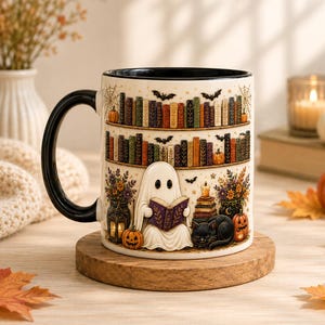 May include: A white ceramic mug with a black handle and interior, featuring a wrap-around illustration of a ghost reading a book, shelves filled with colorful books, bats, pumpkins, a black cat, and floral arrangements in lanterns.