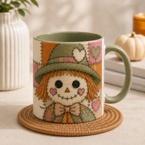 May include: A ceramic mug with a sage green interior and handle, featuring a printed patchwork design of a scarecrow with button eyes, stitched details, and a wide-brimmed hat. The mug displays a light-coloured background with orange, green, and pink accents.