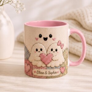 May include: A ceramic mug with a pink interior and handle, featuring a beige speckled exterior with a printed design of two ghosts holding a pink heart, a small ghost above them, and a label at the bottom with the text Ethan & Sophia.