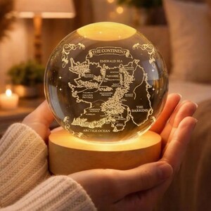 May include: A clear glass globe with a map of a continent etched on its surface, illuminated by a warm light. The globe sits on a wooden base. Text on the globe includes "The Continent," "Emerald Sea," and "Arctic Ocean."
