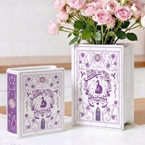 May include: Two white ceramic book-shaped vases with purple accents and the text "The Archer and the Fox." The larger vase holds pink roses. The vases have a decorative floral design.