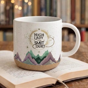 Könnte beinhalten: Weiße Keramik-Tasse mit dem Text "HIGH LADY of the SMUT COURT" in einem kreisförmigen Design. Die Tasse zeigt ein Bergketten-Design in Grün-, Lila- und Brauntönen. Die Tasse steht auf einem offenen Buch.
