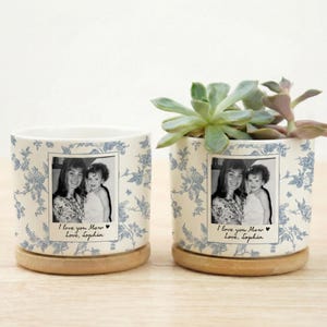 May include: Two white ceramic plant pots with blue floral patterns and wooden bases. Each pot has a black and white photo and the text "I love you Mom, Love, Sophia". One pot contains a succulent plant.