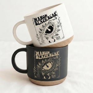 Könnte beinhalten: Zwei Keramikbecher, einer weiß und einer schwarz, übereinander gestapelt. Jeder Becher trägt den Text "MANON BLACKBEAK WORLD TOUR 13" und eine Illustration eines Drachenauges. Der Boden jedes Bechers ist naturfarben.