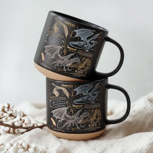 May include: Two black ceramic mugs stacked on top of each other. Each mug features illustrations of dragons, with text including the names "Andarna", "Sgaerl", and "Tairn". The mugs have a tan base and black handles.