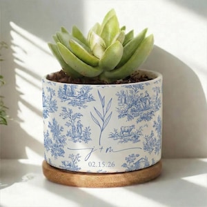 以下が含まれることがあります： 青いトワルドジュイ模様の白いセラミックポットに入った小さな多肉植物。ポットは丸い木の台座の上にあります。ポットには青い葉のデザインと「j + m」のイニシャルと日付「02.15.26」があります。