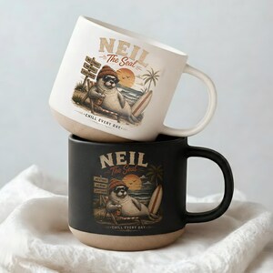 May include: A white ceramic mug stacked on a black ceramic mug. Both mugs feature a graphic of a seal wearing a beanie and sunglasses, sitting on a beach chair with a surfboard, palm tree, and the text Neil The Seal Chill Every Day.
