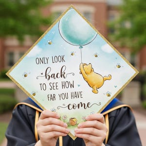 May include: A graduation cap with a gold glitter border, featuring a cartoon illustration of Winnie the Pooh holding a balloon. The cap has the text "Only look back to see how far you have come" with bees and flowers.