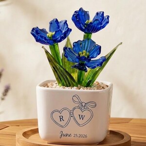 May include: A decorative glass flower arrangement featuring three blue flowers in a white square pot. The pot has two heart-shaped designs with the letters "R" and "W" and the date "June 25.2026". The arrangement sits on a wooden tray.