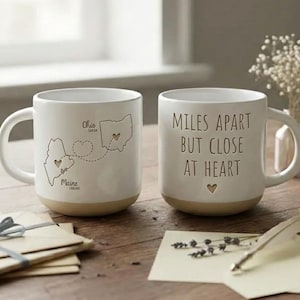 Pode incluir: Duas canecas de cerâmica branca com bases bege. Uma caneca apresenta um mapa de Ohio e Maine com um coração, a outra diz "Miles Apart But Close At Heart". Um pequeno vaso de flores e artigos de papelaria estão sobre a mesa de madeira.