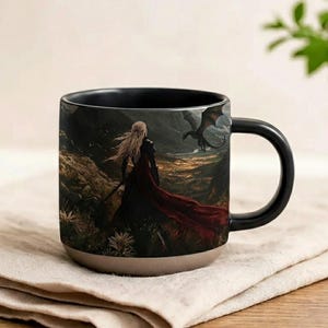 May include: Black ceramic mug featuring fantasy artwork. The design shows a woman with long blonde hair and a red cloak, set against a mountainous backdrop with a dragon. The mug has a black handle and interior, with a beige base.
