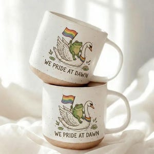 May include: Two speckled off-white ceramic mugs stacked vertically. Each mug features a green frog riding a white swan, holding a rainbow flag. A small rainbow collar is on the swan. The text WE PRIDE AT DAWN is printed below the illustration.