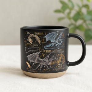 May include: Black ceramic mug with a tan base, featuring illustrations of dragons and text. The dragons are in various poses with different wing designs. The mug has a black handle and the text includes the names "Andarna", "Sgaeyl", and "Tairn".