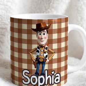 May include: A white ceramic mug featuring a brown and cream gingham pattern background. The mug displays a graphic of a cowboy character wearing a brown hat, yellow shirt, and cow-print vest. The name Sophia is printed in large white letters.