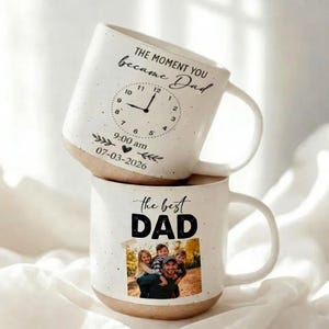 Op de afbeelding: Twee witte keramische mokken op elkaar gestapeld. De bovenste mok heeft de tekst "THE MOMENT YOU became Dad" en een klokontwerp. De onderste mok zegt "the best DAD" met een familiefoto.