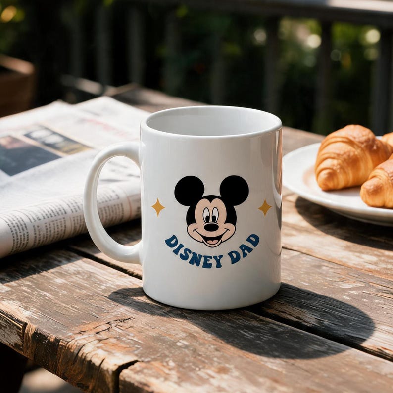 Puede incluir: Una taza de cer&aacute;mica blanca con un gr&aacute;fico del rostro de Mickey Mouse centrado sobre el texto azul "DISNEY DAD", con dos peque&ntilde;as estrellas amarillas de cuatro puntas a ambos lados de las orejas del personaje y un asa curva.