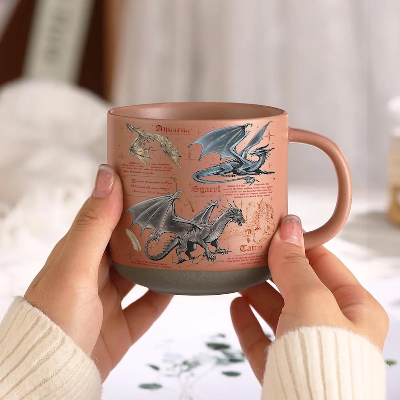May include: A ceramic mug with a pinkish-brown exterior and a gray base. The mug features illustrations of dragons with the names "Anvarn", "Sgaerl", and "Tairn" printed on the side. The mug has a large handle.