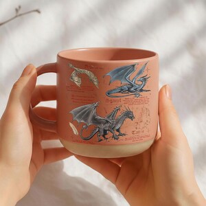 May include: A ceramic mug with a peach-colored upper half and a beige base. The mug features illustrations of three different dragons with detailed wings and scales. The word "Tain" is visible on the mug.