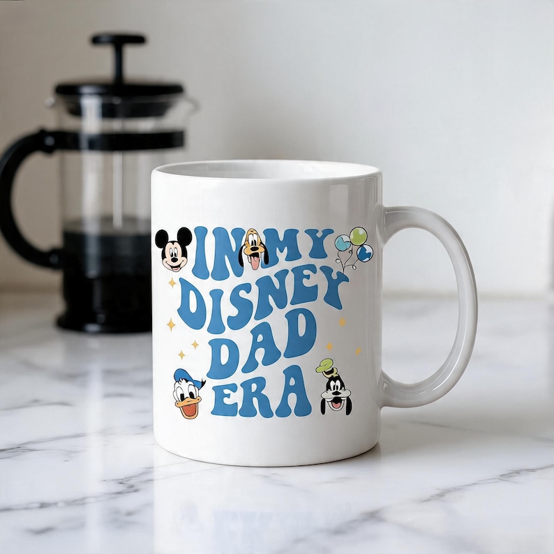 Puede incluir: Una taza de cer&aacute;mica blanca con el texto azul "IN MY DISNEY DAD ERA" en una fuente ondulada. El dise&ntilde;o incluye peque&ntilde;as ilustraciones de Mickey Mouse, Goofy, el Pato Donald y Pluto, junto con globos coloridos y detalles de estrellas amarillas.