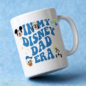 Puede incluir: Una taza de cer&aacute;mica blanca con el texto azul IN MY DISNEY DAD ERA. El dise&ntilde;o incluye ilustraciones coloridas de Mickey Mouse, Pluto, el Pato Donald y Goofy, junto con peque&ntilde;os detalles de estrellas amarillas y tres globos.