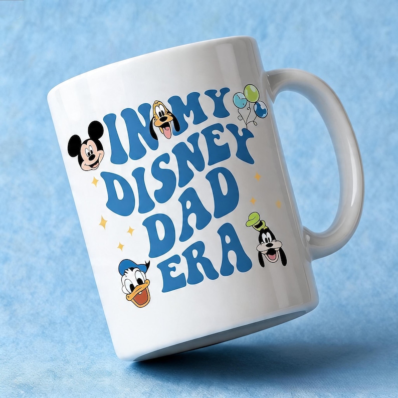 Puede incluir: Una taza de cer&aacute;mica blanca con el texto azul IN MY DISNEY DAD ERA. El dise&ntilde;o incluye ilustraciones coloridas de Mickey Mouse, Pluto, el Pato Donald y Goofy, junto con peque&ntilde;os detalles de estrellas amarillas y tres globos.