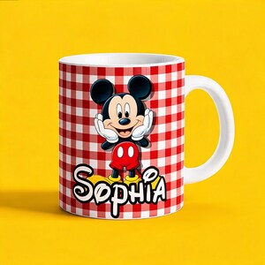 K&ouml;nnte beinhalten: Ein wei&szlig;er Keramikbecher mit einem rot-wei&szlig;en Karomuster. Auf der Vorderseite befindet sich eine Grafik von Mickey Mouse mit den H&auml;nden an den Wangen, platziert &uuml;ber dem Namen Sophia in einer fetten, wei&szlig;-schwarz umrandeten Schreibschrift.