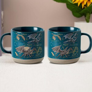 May include: Two teal ceramic mugs with a dragon design. Each mug features a dark teal interior, a speckled beige base, and a wraparound illustration of dragons with text. The mugs have a curved handle.