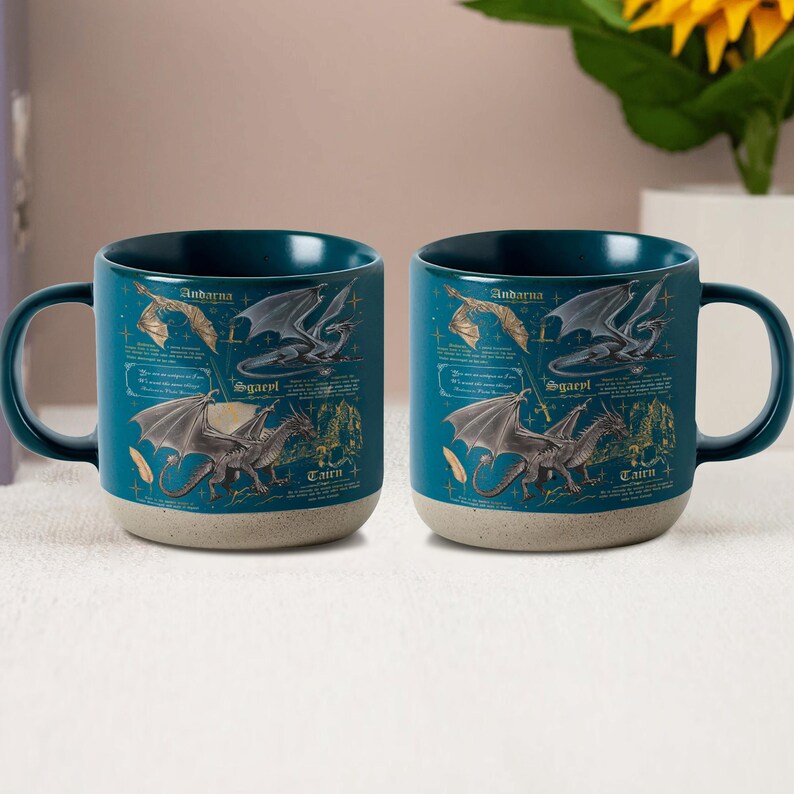 May include: Two teal ceramic mugs with a dragon design. Each mug features a dark teal interior, a speckled beige base, and a wraparound illustration of dragons with text. The mugs have a curved handle.