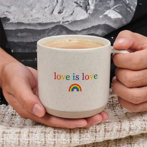 May include: A speckled off-white ceramic mug featuring the text love is love printed in multicolored letters with a small rainbow icon underneath. The mug has a smooth handle and a solid base, containing a light brown liquid inside the vessel.