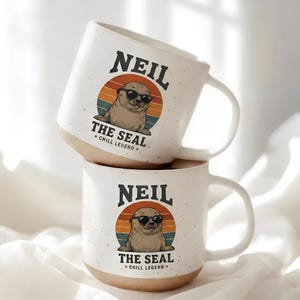May include: Two off-white ceramic mugs with a speckled finish. Each mug features a graphic of a brown seal wearing black sunglasses centered within a sunset-colored circle. The text reads NEIL THE SEAL and CHILL LEGEND with two small red stars.