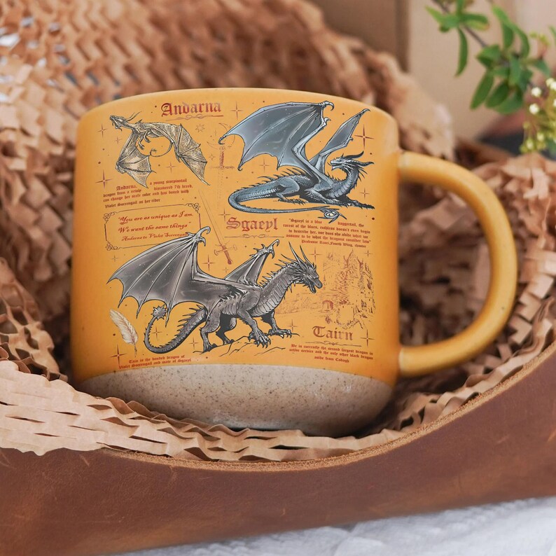 May include: Mustard yellow ceramic mug featuring illustrations of dragons with the names Andarna, Sgaeyl, and Cairn. The mug has a tan base and a matching yellow handle. The mug is presented in a brown leather container with brown paper.
