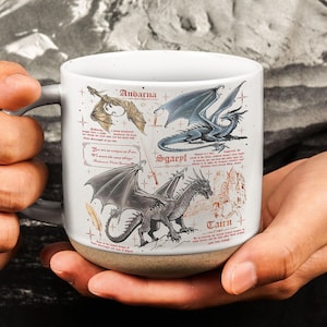 May include: White ceramic mug featuring illustrations of dragons with names like Andarna, Sgaeyl, and Tairn. The mug has a gray handle and a tan base. The dragons are in various colors, including gold, blue, and gray.