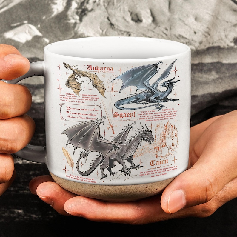 May include: White ceramic mug featuring illustrations of dragons with names like Andarna, Sgaeyl, and Tairn. The mug has a gray handle and a tan base. The dragons are in various colors, including gold, blue, and gray.