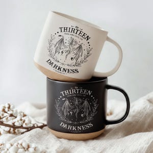May include: Two ceramic mugs, one white and one black, stacked. Each mug features a circular design with the text "the THIRTEEN FROM NOW UNTIL THE DARKNESS CLAIMS US" and a bat-like creature illustration. The bottom of each mug is a natural tan color.
