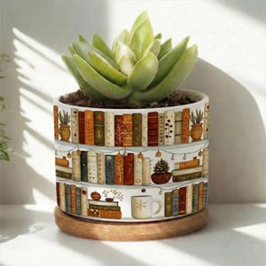 Op de afbeelding: Witte keramische bloempot met een ontwerp van boekenkasten vol boeken, lichtslingers en een koffiekopje. De pot bevat een vetplant en staat op een ronde houten basis. Het ontwerp toont kleurrijke boeken.