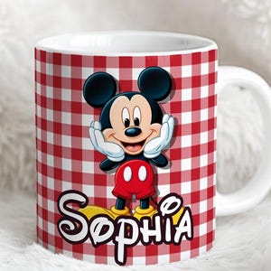 K&ouml;nnte beinhalten: Ein wei&szlig;er Keramikbecher mit einem rot-wei&szlig;en Vichy-Karo-Muster. Der Becher zeigt eine Grafik von Mickey Mouse mit den H&auml;nden an den Wangen, platziert &uuml;ber dem Namen Sophia, der in einer stilisierten wei&szlig;en Schrift mit schwarzem Rand steht.