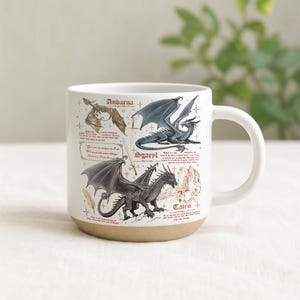 May include: White ceramic mug with a tan base, featuring illustrations of three dragons with names: Andarna, Sgaeyl, and Tairn. The mug has a curved handle and includes text descriptions of each dragon. The background is a soft, neutral color.