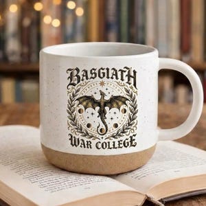 Könnte beinhalten: Eine weiße Keramiktasse mit braunem Boden, die eine detaillierte Illustration eines Drachen in einem kreisförmigen Design zeigt. Der Text "BASGIATH WAR COLLEGE" ist oberhalb und unterhalb der Drachenillustration zu sehen.