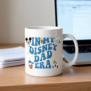 Puede incluir: Una taza de cer&aacute;mica blanca con texto azul que dice IN MY DISNEY DAD ERA. El dise&ntilde;o incluye peque&ntilde;as ilustraciones coloridas de Mickey Mouse, Goofy, el Pato Donald y un grupo de globos, acentuado por diminutas estrellas amarillas dispersas.