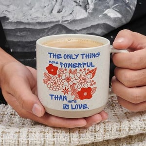 May include: A speckled ceramic mug with a floral design and the text "THE ONLY THING MORE POWERFUL THAN HATE IS LOVE" in blue. The mug is filled with a light brown liquid.